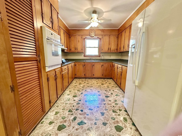 kitchen with a ceiling fan, a sink, dark countertops, white appliances, and brown cabinetry