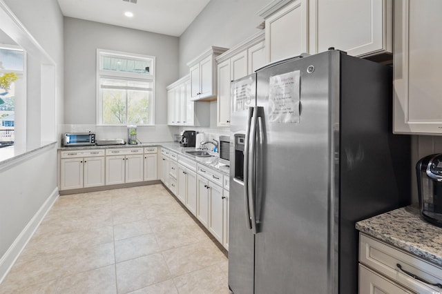 kitchen featuring a sink, light tile patterned flooring, stainless steel fridge with ice dispenser, baseboards, and light stone countertops