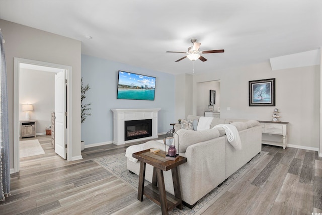 living area with ceiling fan, light wood-style flooring, baseboards, and a glass covered fireplace