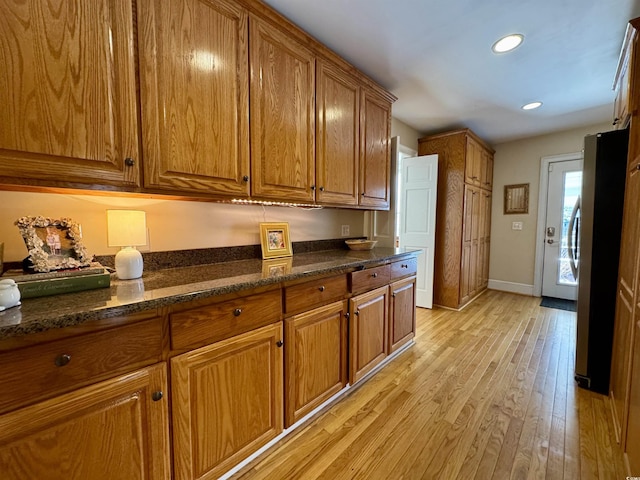 kitchen with baseboards, light wood-type flooring, brown cabinets, dark stone countertops, and freestanding refrigerator