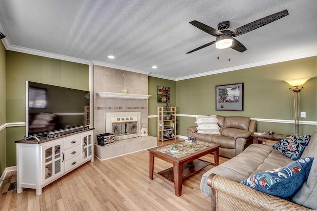 living area featuring ceiling fan, a tiled fireplace, ornamental molding, light wood-style floors, and a textured ceiling