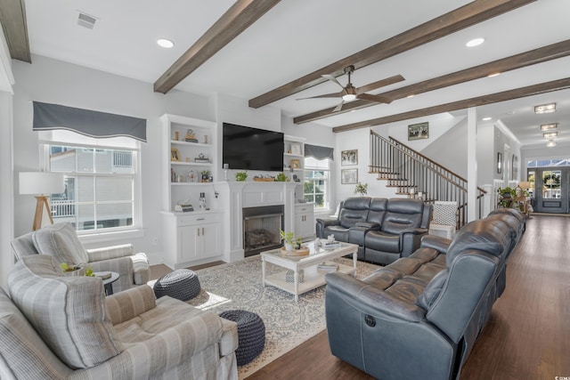 living area with stairway, a fireplace with flush hearth, beamed ceiling, dark wood-style floors, and a ceiling fan