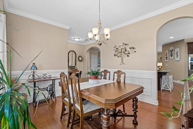 dining area with wood finished floors, a wainscoted wall, arched walkways, ornamental molding, and a notable chandelier