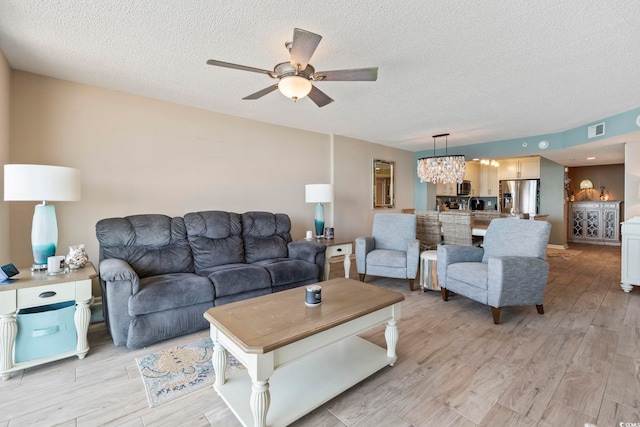 living room featuring ceiling fan with notable chandelier, visible vents, light wood finished floors, and a textured ceiling