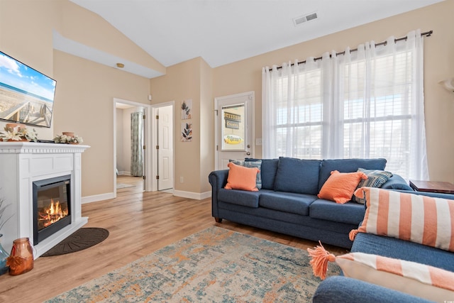 living room featuring baseboards, visible vents, lofted ceiling, light wood-style flooring, and a glass covered fireplace