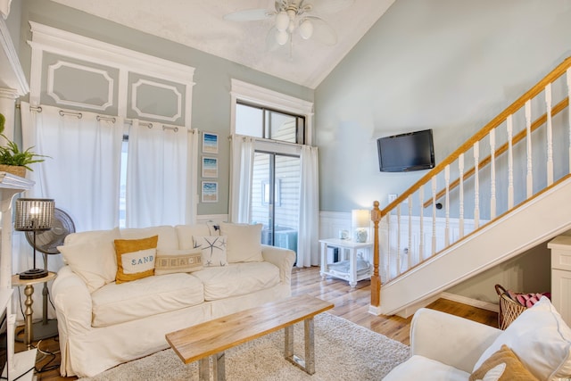 living room with ceiling fan, stairway, a wainscoted wall, light wood-type flooring, and high vaulted ceiling