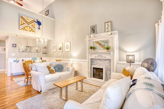 living room featuring a brick fireplace, a wainscoted wall, ceiling fan with notable chandelier, a towering ceiling, and wood finished floors