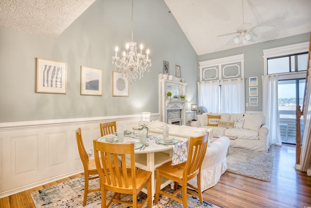 dining area with wood finished floors, ceiling fan with notable chandelier, a wainscoted wall, and high vaulted ceiling