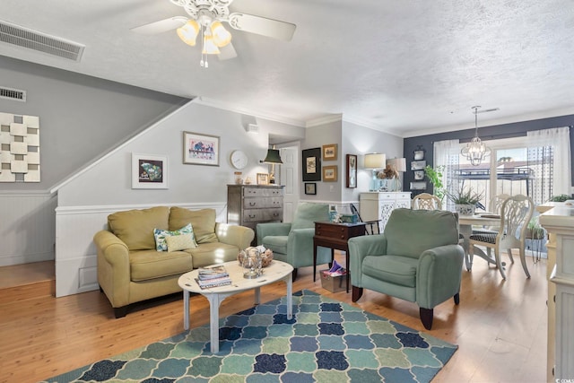 living room featuring visible vents, ornamental molding, light wood-style flooring, ceiling fan with notable chandelier, and a textured ceiling