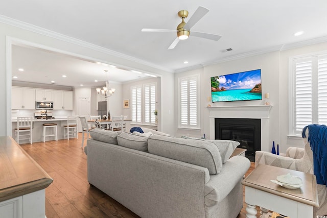 living room featuring visible vents, ornamental molding, ceiling fan with notable chandelier, light wood-style floors, and a glass covered fireplace