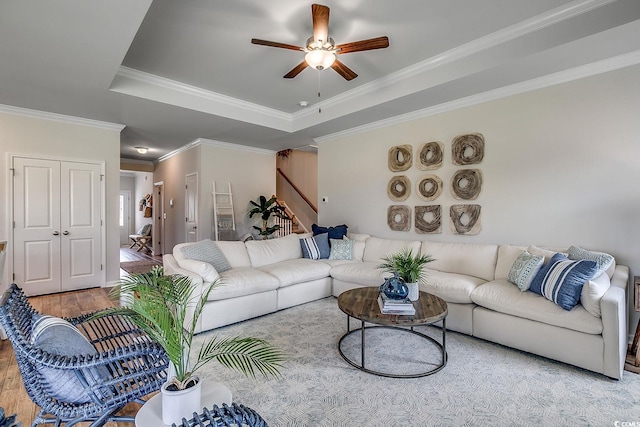 living area with crown molding, wood finished floors, ceiling fan, stairway, and a raised ceiling