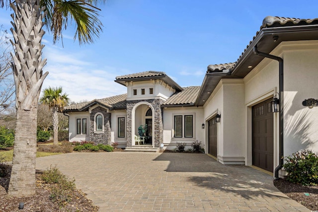 mediterranean / spanish-style home featuring a tiled roof, stone siding, a garage, stucco siding, and decorative driveway