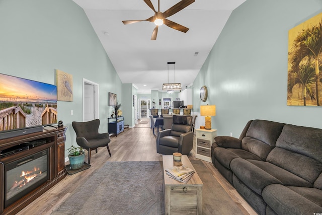 living area with ceiling fan, high vaulted ceiling, a glass covered fireplace, and wood finished floors