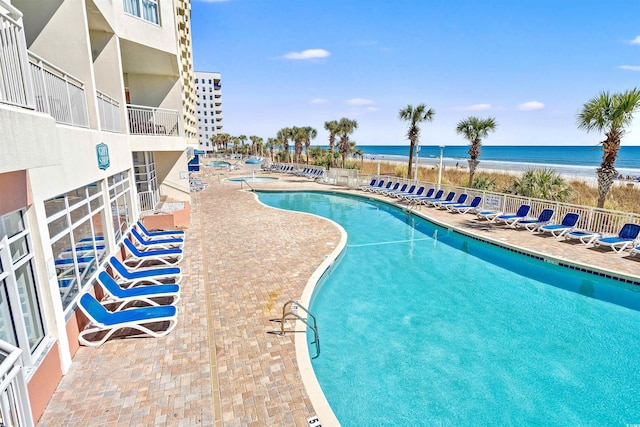 pool with a patio, a water view, fence, and a beach view