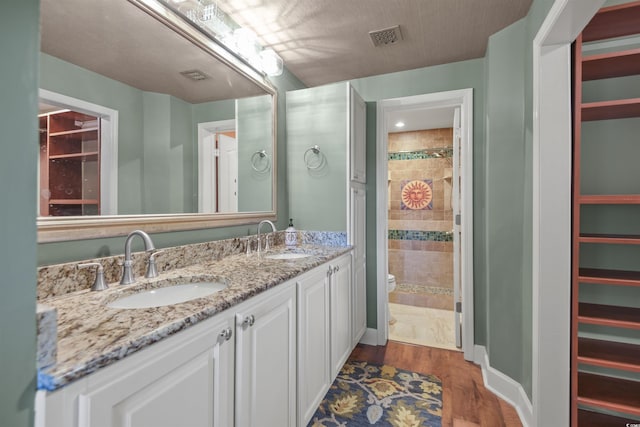 full bathroom with toilet, wood finished floors, tiled shower, double vanity, and baseboards