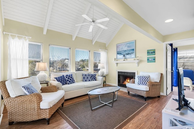 living area featuring ceiling fan, vaulted ceiling with beams, a warm lit fireplace, and wood finished floors