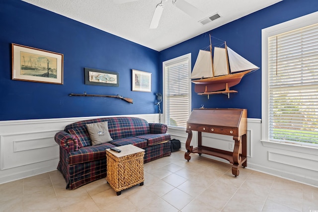living area featuring a ceiling fan, a textured ceiling, visible vents, a wainscoted wall, and tile patterned floors