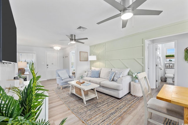 living room with ceiling fan, a decorative wall, light wood finished floors, and visible vents