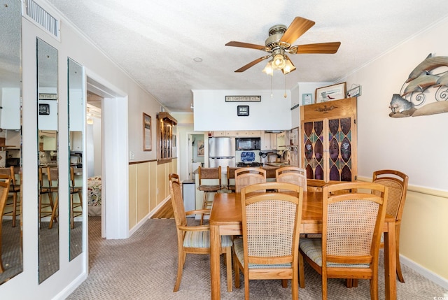 dining area with light carpet, ceiling fan, ornamental molding, and a wainscoted wall