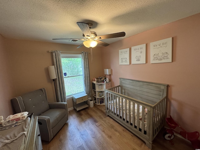 bedroom featuring wood finished floors, a textured ceiling, a crib, and ceiling fan