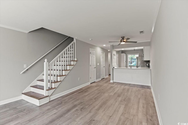 unfurnished living room with a ceiling fan, visible vents, light wood-style floors, ornamental molding, and stairs