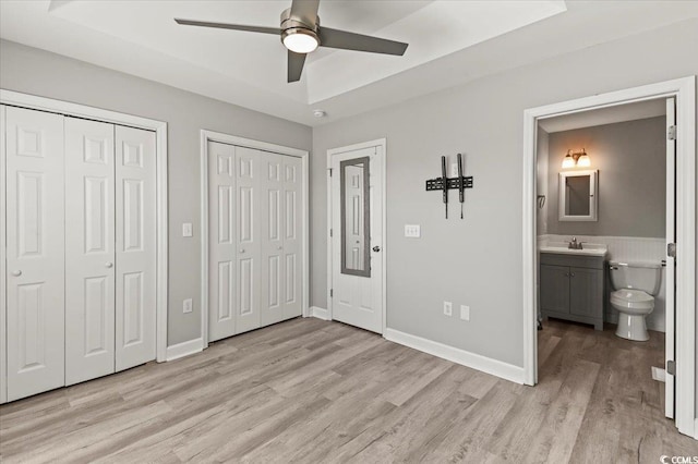 unfurnished bedroom with two closets, baseboards, light wood-type flooring, connected bathroom, and a tray ceiling