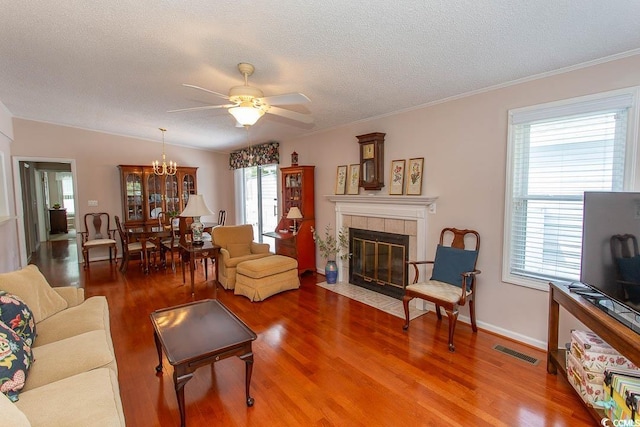 living room featuring wood finished floors, a textured ceiling, ceiling fan with notable chandelier, a tiled fireplace, and visible vents