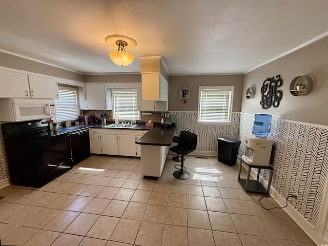 kitchen with black appliances, a peninsula, dark countertops, a wainscoted wall, and healthy amount of natural light
