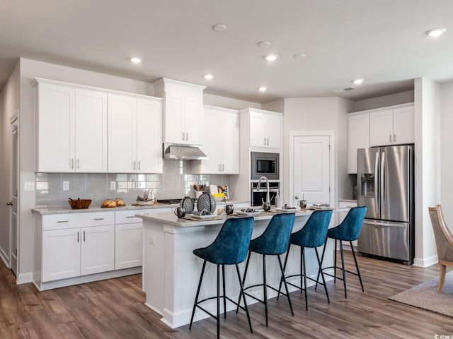 kitchen with dark wood-type flooring, under cabinet range hood, a center island with sink, and stainless steel appliances