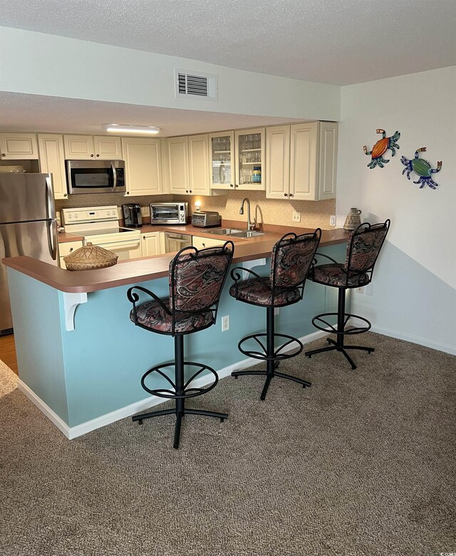 kitchen featuring stainless steel appliances, a kitchen bar, glass insert cabinets, cream cabinetry, and a peninsula
