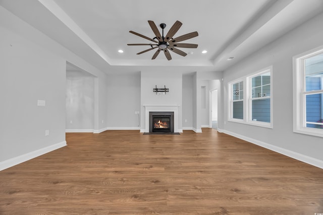 unfurnished living room with a ceiling fan, a fireplace with flush hearth, wood finished floors, baseboards, and a tray ceiling