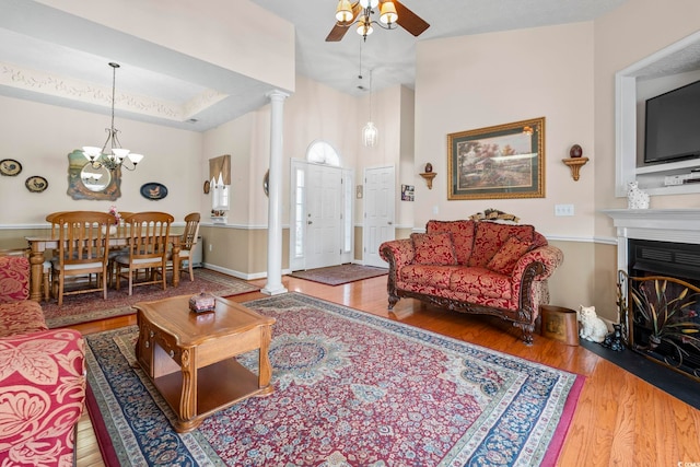 living area featuring a glass covered fireplace, ceiling fan with notable chandelier, ornate columns, and wood finished floors