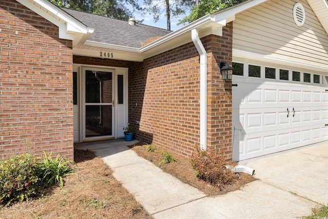view of exterior entry featuring brick siding, driveway, a shingled roof, and a garage