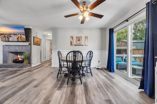 dining space featuring ceiling fan, wainscoting, a brick fireplace, a textured ceiling, and wood finished floors