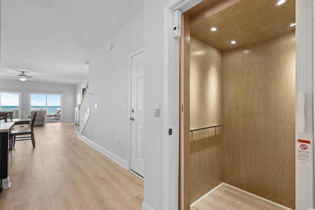 corridor featuring light wood finished floors, elevator, recessed lighting, baseboards, and stairway