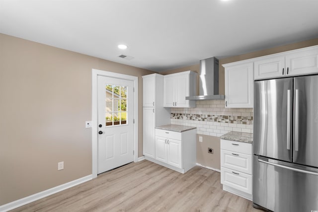 kitchen with light wood finished floors, freestanding refrigerator, wall chimney exhaust hood, white cabinets, and backsplash