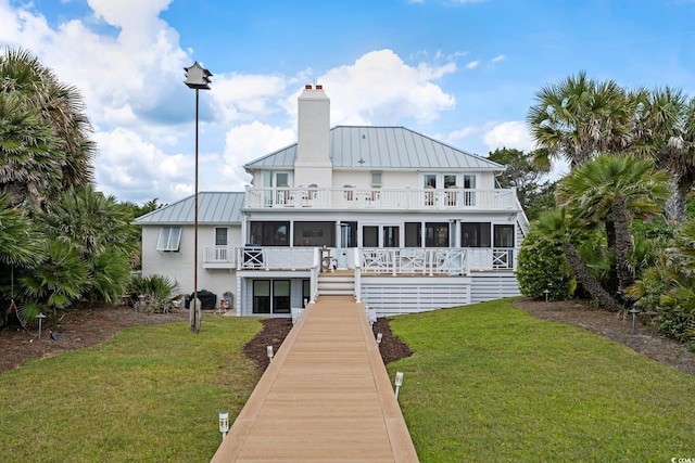 rear view of property featuring a balcony, a chimney, a standing seam roof, a sunroom, and a lawn