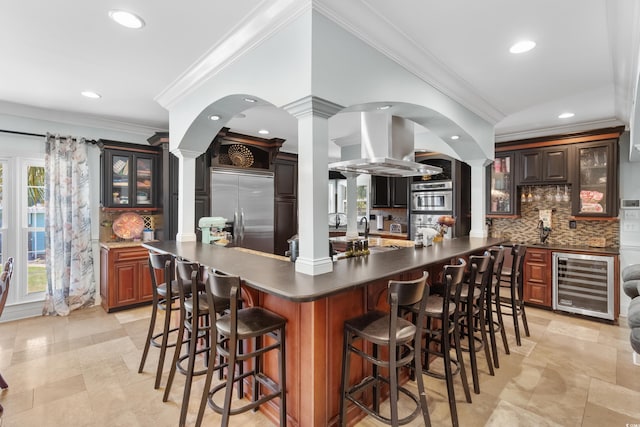 kitchen featuring island exhaust hood, ornate columns, a breakfast bar, appliances with stainless steel finishes, and wine cooler