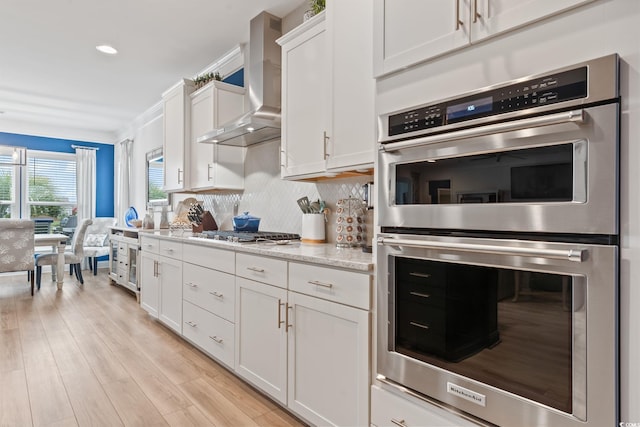 kitchen featuring wall chimney range hood, light wood-type flooring, white cabinets, backsplash, and stainless steel appliances