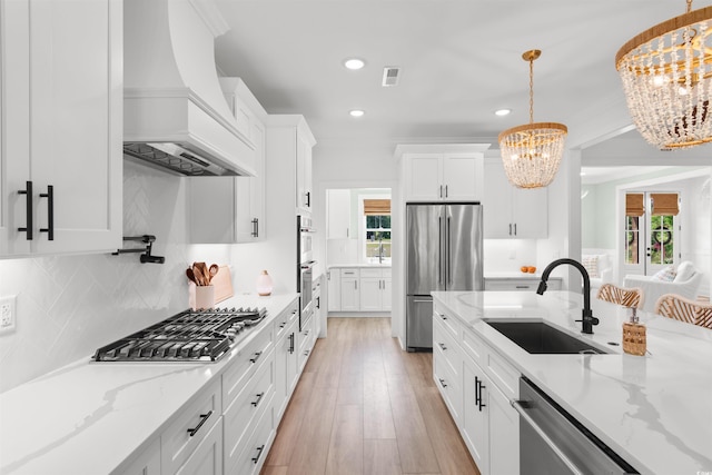 kitchen with a sink, custom exhaust hood, a notable chandelier, stainless steel appliances, and white cabinetry