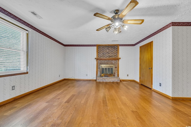 unfurnished living room with wood finished floors, ceiling fan, a brick fireplace, crown molding, and wallpapered walls