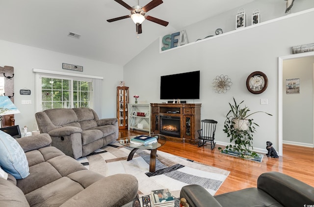 living room with ceiling fan, a lit fireplace, visible vents, high vaulted ceiling, and light wood-style floors