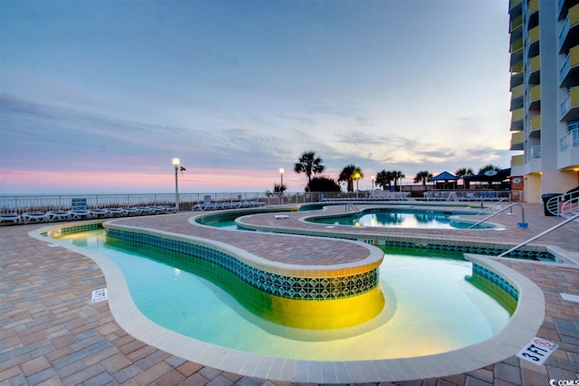 pool at dusk featuring a hot tub, a community pool, a patio, and fence