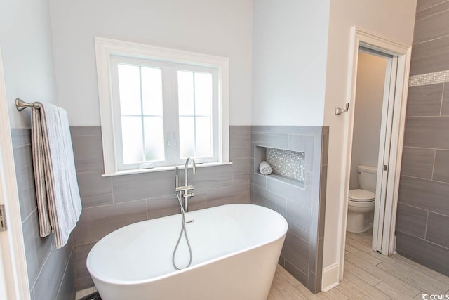 full bathroom with wood finished floors, toilet, tile walls, a wainscoted wall, and a soaking tub