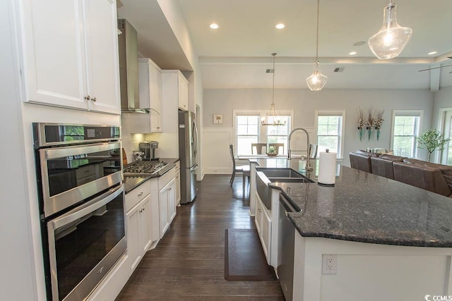 kitchen featuring a large island with sink, white cabinets, open floor plan, stainless steel appliances, and wall chimney exhaust hood