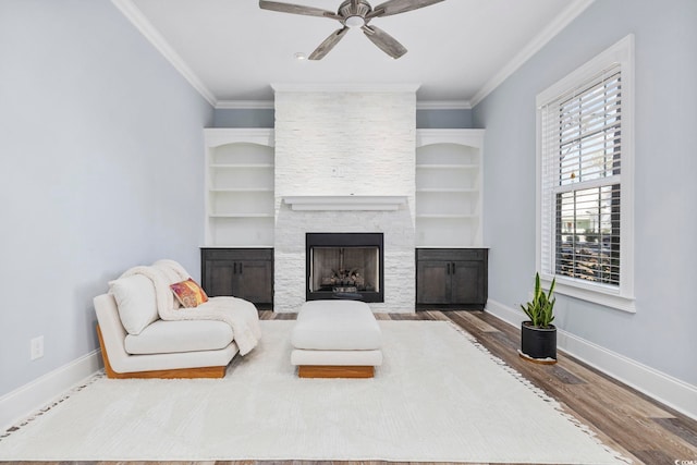 living room featuring a stone fireplace, a ceiling fan, baseboards, wood finished floors, and ornamental molding
