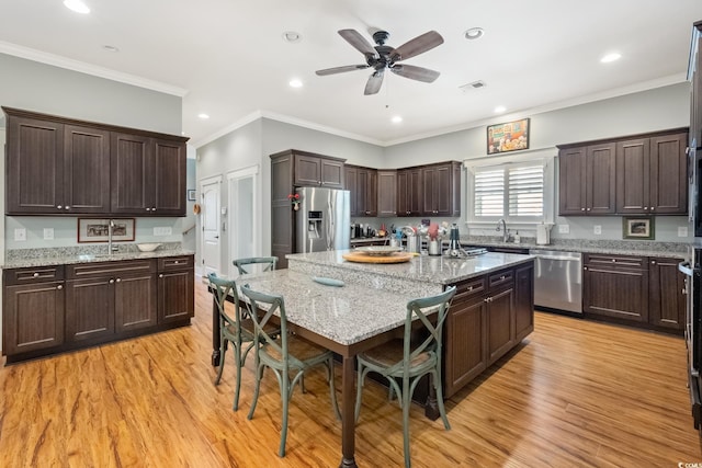 kitchen with light wood finished floors, a kitchen bar, an island with sink, dark brown cabinetry, and appliances with stainless steel finishes