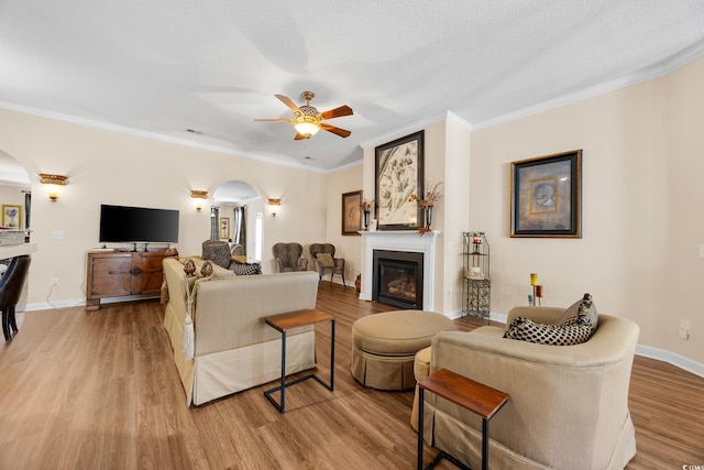 living room with arched walkways, wood finished floors, a glass covered fireplace, crown molding, and ceiling fan