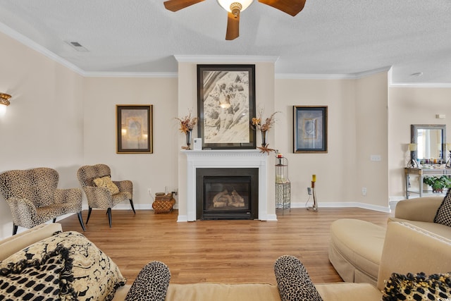 living area featuring light wood-type flooring, crown molding, a textured ceiling, a glass covered fireplace, and ceiling fan