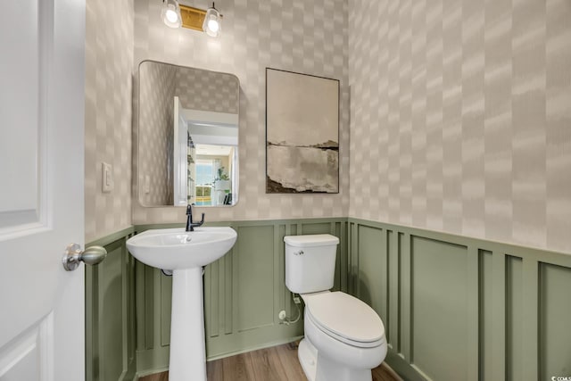half bath with wallpapered walls, toilet, wainscoting, a sink, and wood finished floors